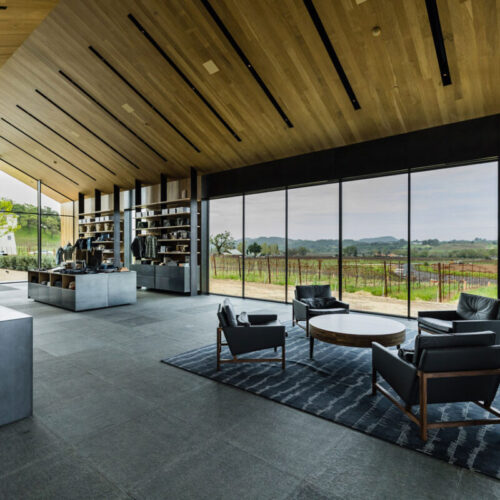 Modern open-plan lounge with wood plank ceiling, black linear recessed lights, and floor-to-ceiling glass overlooking vineyards.