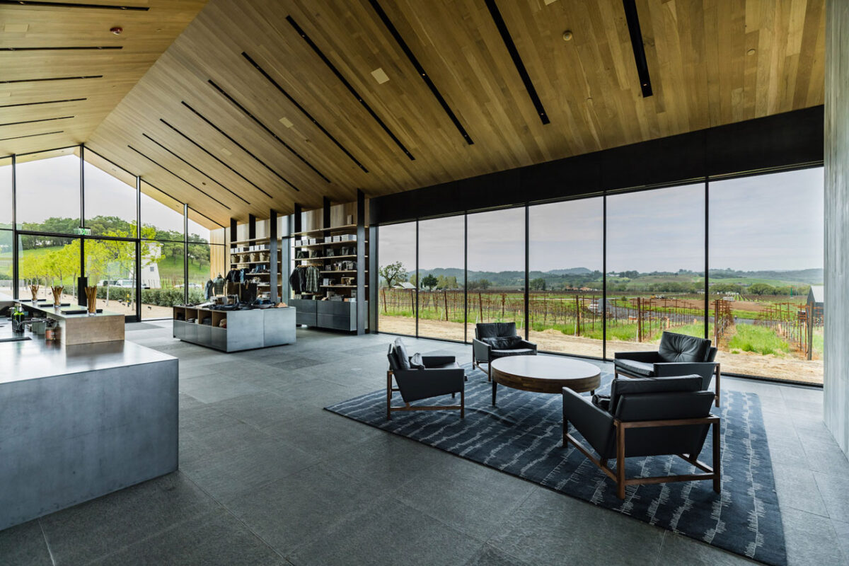 Modern open-plan lounge with wood plank ceiling, black linear recessed lights, and floor-to-ceiling glass overlooking vineyards.