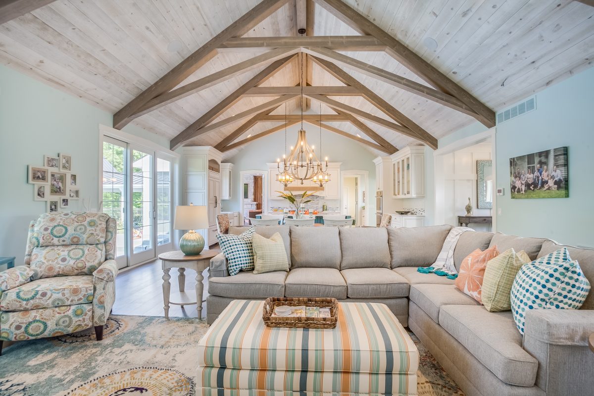 Open-concept living room with gray sectional, striped ottoman, and whitewashed wood plank vaulted ceiling with exposed timber trusses, looking into a white kitchen with chandelier.