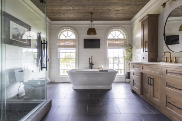 Primary bathroom with white shiplap walls, weathered wood plank ceiling, arched windows, and a freestanding soaking tub between the windows.