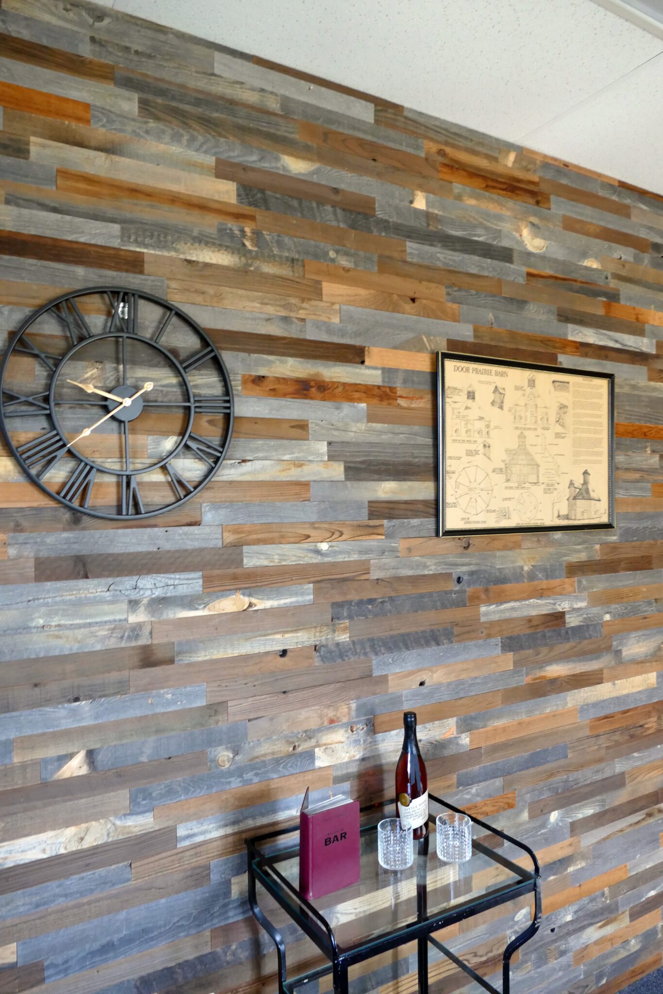 Mixed-tone reclaimed wood plank accent wall with a large black metal wall clock, framed vintage print, and a small bar cart.