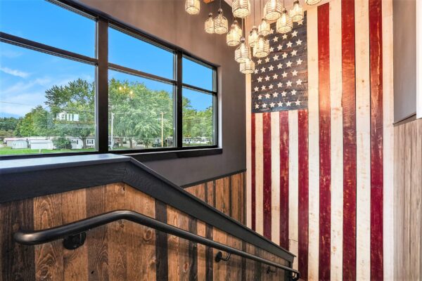 Stairwell landing with reclaimed wood wainscoting, large wooden American flag wall art, pendant lights, and black-framed windows.