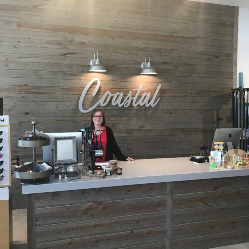 Retail checkout counter with a weathered gray wood plank wall and a large “Coastal” sign lit by two wall sconces.