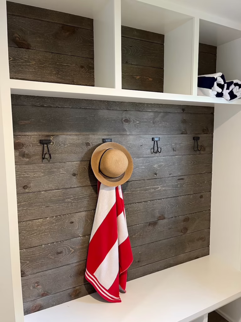 Mudroom cubby with white built-in shelves, weathered gray wood plank back panel, black coat hooks, and a straw hat with a red-and-white towel.