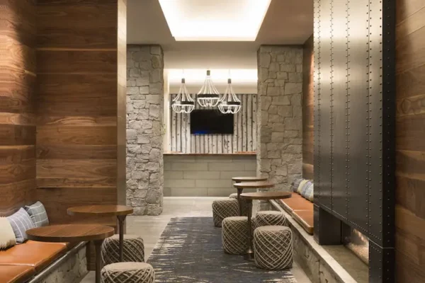 Modern lounge with warm wood plank wall paneling, stone columns, built-in bench seating, and woven ottomans under recessed ceiling lighting.